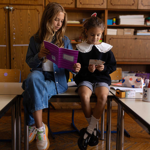 Zwei Mädchen sitzen in einem Klassenzimmer auf einem Tisch, eine hält ein Heft, die andere eine Gesprächskarte
